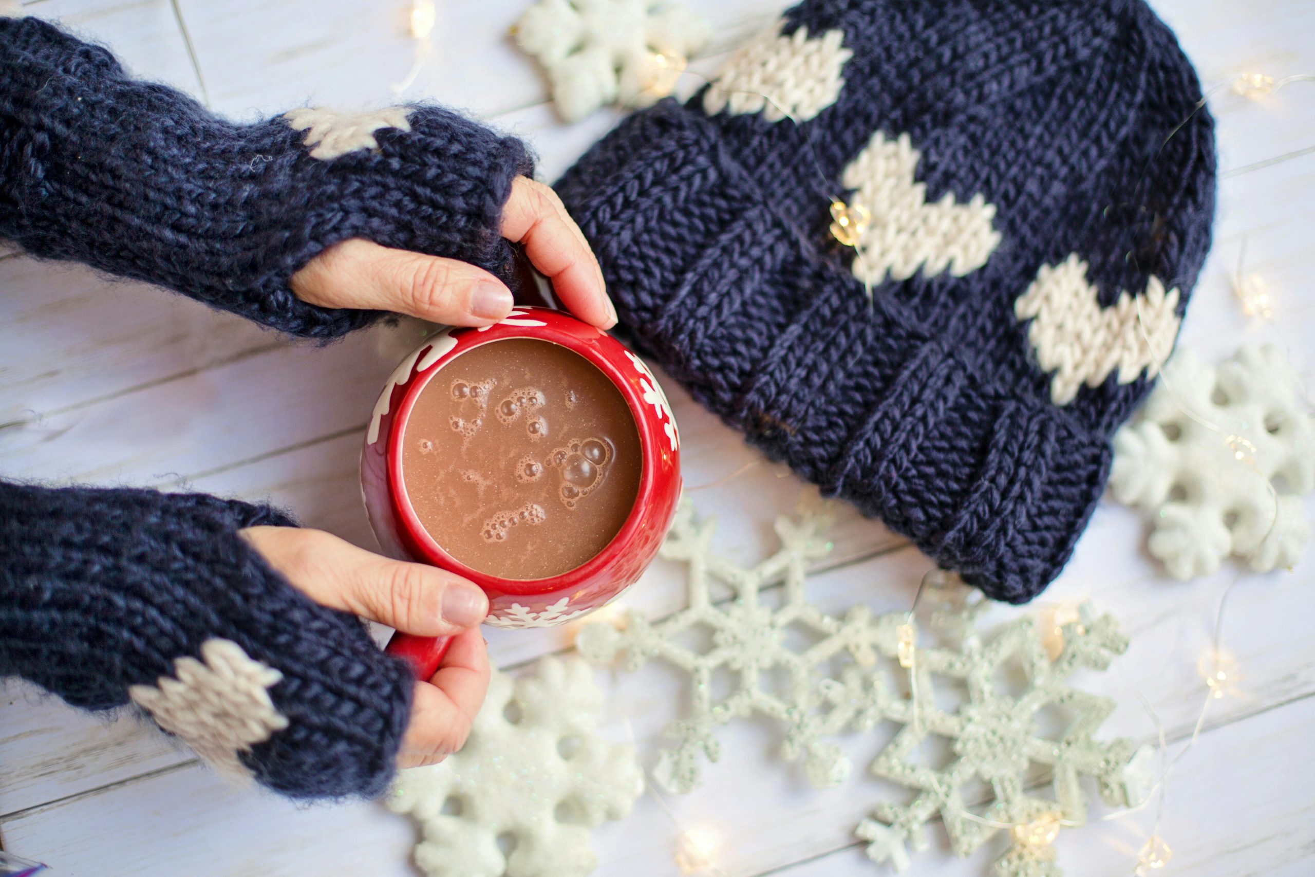 Warm knitted mittens holding hot chocolate during winter with beanie and snowflakes.